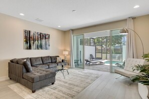 Ein modernes Wohnzimmer mit einem Ecksofa, einer Glastür, die auf einen Balkon führt, und Blick auf einen Pool.