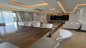 A modern living room with a large flat-screen TV, a dining table, and several sofas.