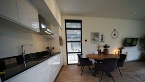 Una cocina moderna con armarios blancos y encimera negra, un comedor con mesa de madera y sillas negras, y un ventanal con persianas.