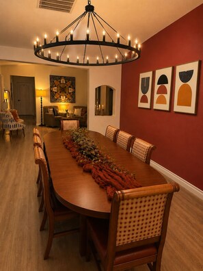 Une salle à manger avec une longue table en bois, des chaises, un lustre, des œuvres d’art accrochées au mur et une porte au fond.