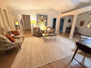 A living room with a large rug, a coffee table, and a sofa. There is a painting on the wall and a lamp on the table.