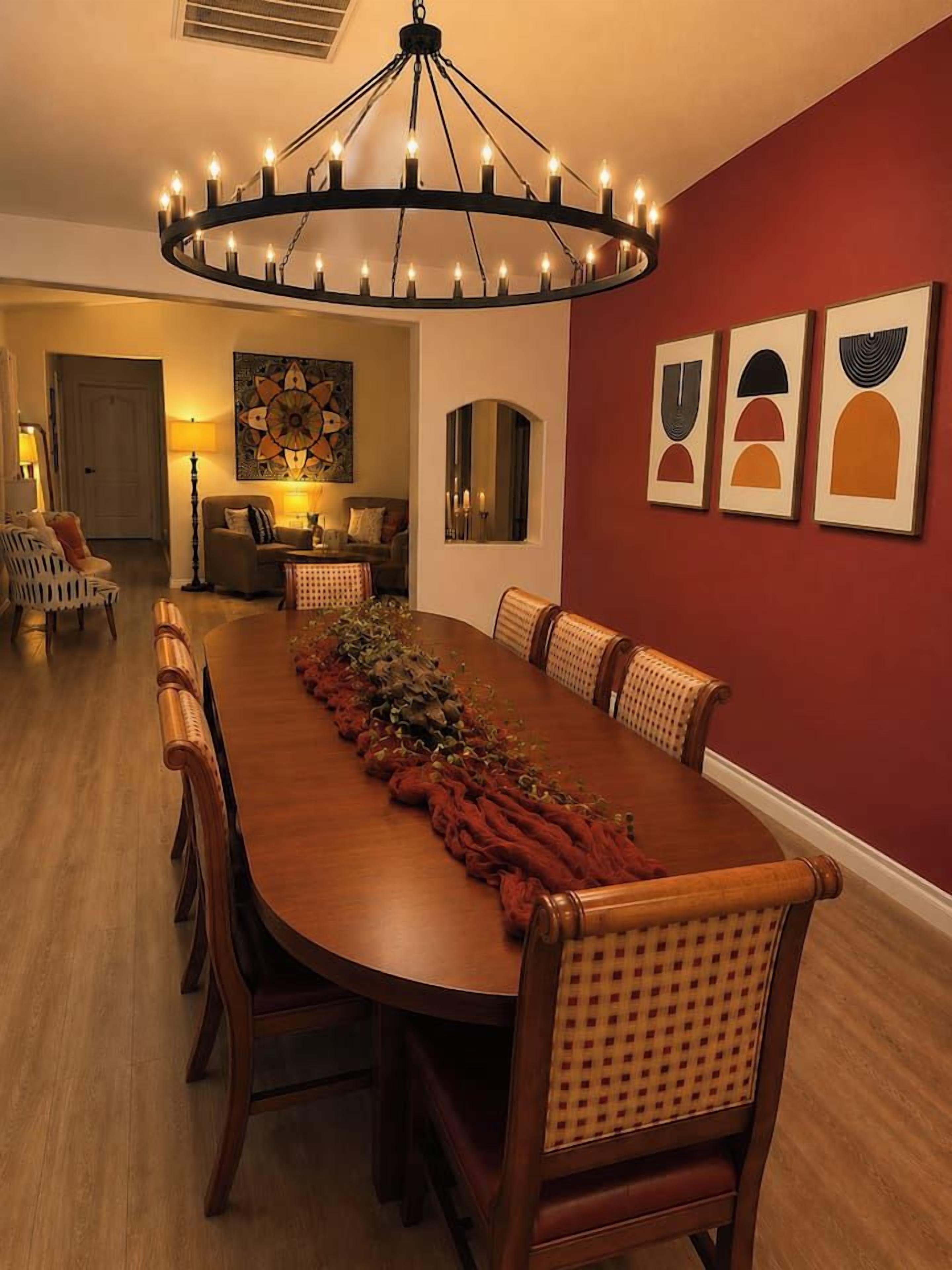 Uma sala de jantar com uma longa mesa de madeira, cadeiras, um candelabro e obras de arte nas paredes.