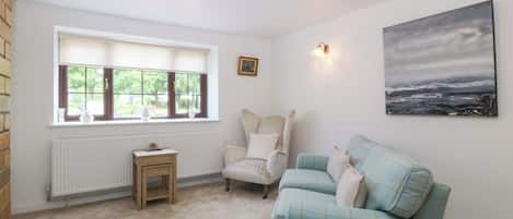 Una habitación con ventana, radiador, una mesita, una silla, un sofá y un cuadro en la pared.