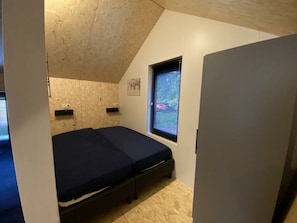 Ein kleines Zimmer mit einem Einzelbett, schrägen Decken, einem Fenster mit Jalousien und einer Tür.