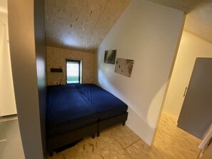 Ein kleines Zimmer mit einem Einzelbett, geneigter Decke, einem Fenster mit Jalousien und einem an der Wand hängenden Bild.