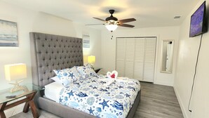 A bedroom with a tufted headboard, a bed with a nautical-themed bedspread, a glass side table with a lamp, a ceiling fan, and a television mounted on the wall. - 2-bedroom house in fantastic Hudson with cool AC breeze (Hudson)