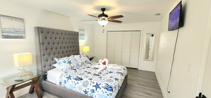 Ein Schlafzimmer mit einer gepolsterten Kopfteile, einem Bett mit einem marineblauen Bettbezug, einem Glastische mit einer Lampe, einem Deckenventilator und einem an der Wand montierten Fernseher.