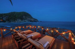 A wooden deck with lounge chairs and string lights, overlooking a coastal area with a rocky outcrop and ocean. - Bichu Boutique (Puerto Ángel)