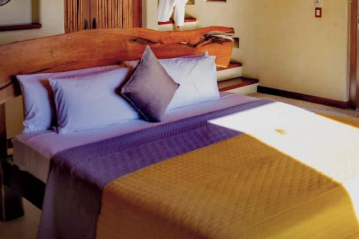 Un lit avec une tête de lit en bois, une couvre-lit aux couleurs violettes et jaunes, et un oreiller blanc.