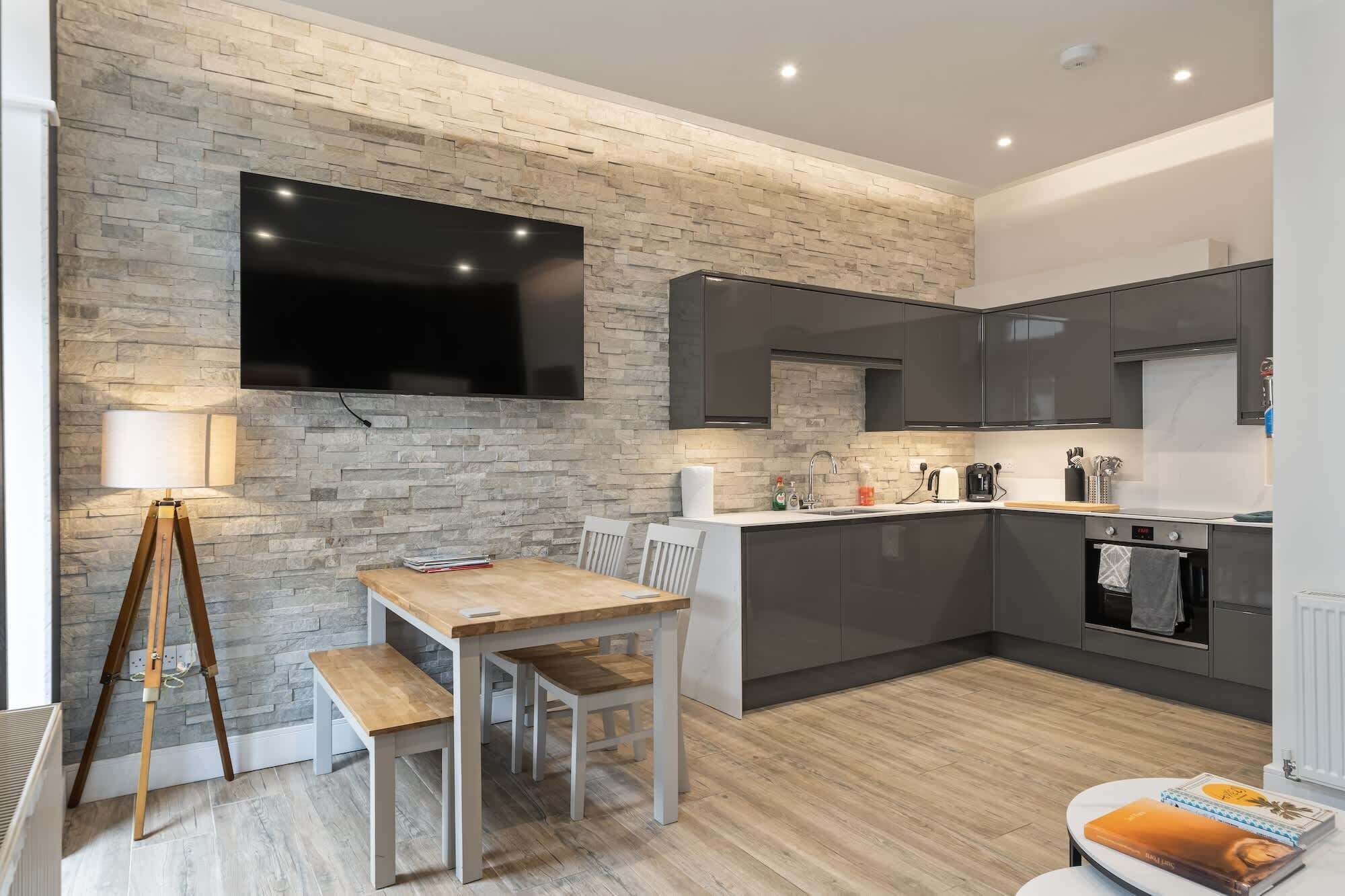 A modern kitchen with a stone wall, a flat-screen TV, and a dining area with a wooden table and chairs.