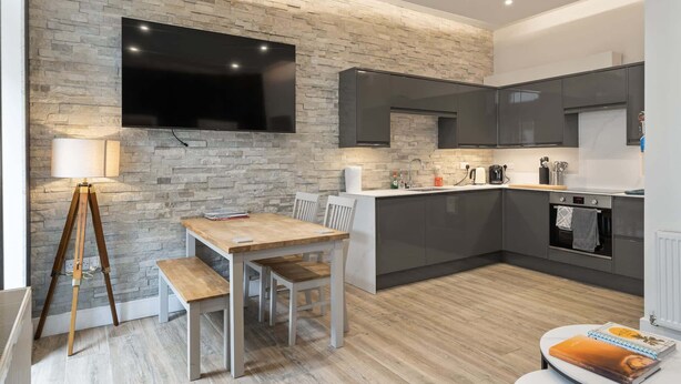 Ett modernt kök med en vägg av natursten, en platt-TV och en matplats med ett bord och stolar i trä.