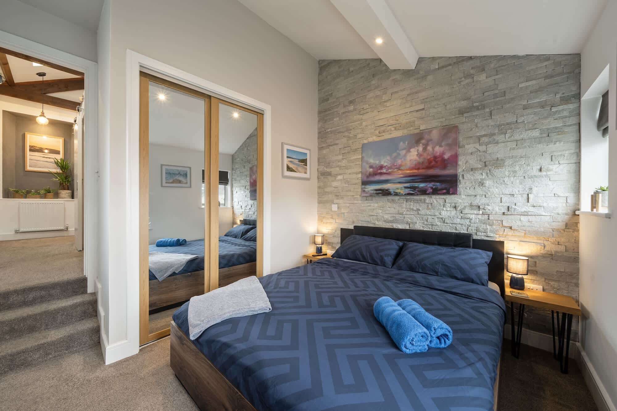 A bedroom with a large bed, a stone wall, a painting, and a view into another room.