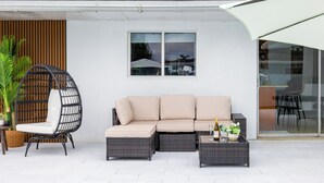 Zona junto a la piscina con un sofá de mimbre, una silla colgante y una pequeña mesa con una botella y flores.