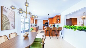 Una cocina moderna con armarios de madera, una zona de comedor con mesa de madera y sillas tapizadas en verde, y una lámpara colgante.