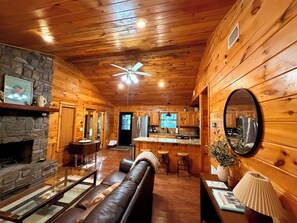 A cozy cabin interior with a stone fireplace, leather sofa, wooden ceiling, and a kitchen area. - Cloud Nine Cabin Private 2 bed 2 bath 2 Living Hot Tub Outdoor Fireplace Sunroom (Broken Bow)