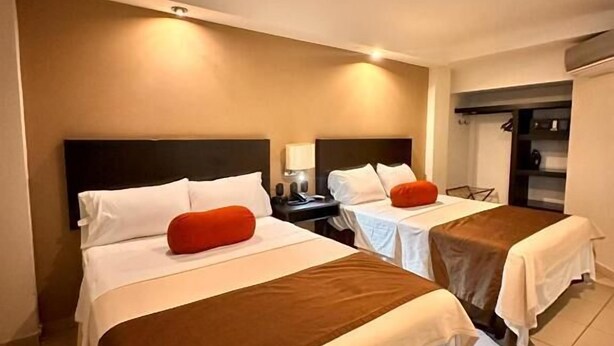 Quarto de hotel com duas camas, cada uma com travesseiro e cobertor. Apresenta uma mesinha de cabeceira com luminária e uma pequena mesa com cadeira. O piso é revestido com azulejos e há uma luminária no teto.