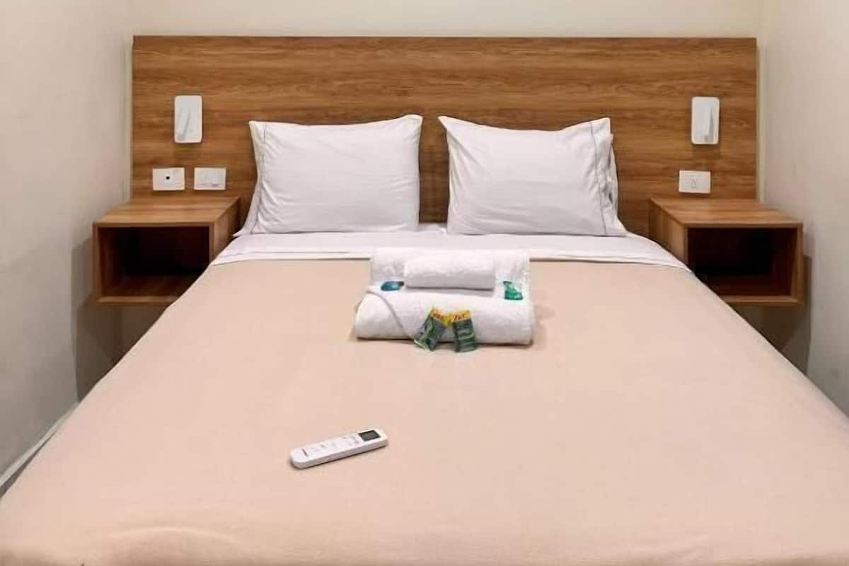 A hotel room with a wooden headboard, two white pillows, a beige bedspread, a remote control, and a neatly folded towel on the bed.