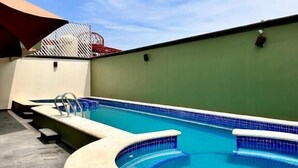 Una piscina con borde curvo, un toldo rojo y un cielo azul despejado.