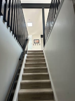 Ein mit Teppich ausgelegter Treppenaufgang mit schwarzen Geländern, der zu einem Raum mit einem gerahmten Bild und einem Lichtschalter an der Wand führt.