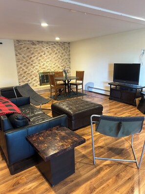 Ein Wohnzimmer mit einer Couch, einem Hocker, einem Couchtisch, einem Fernseher und einem Essbereich mit Stühlen und einem Tisch.