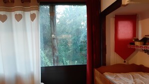 Une chambre à coucher avec un lit, une petite table ornée d’un objet décoratif, une fenêtre donnant sur des arbres et un mur rouge.