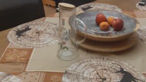 Un coin repas avec une table dressée pour deux personnes, un téléviseur en arrière-plan et une cuisine visible à travers une porte ouverte.