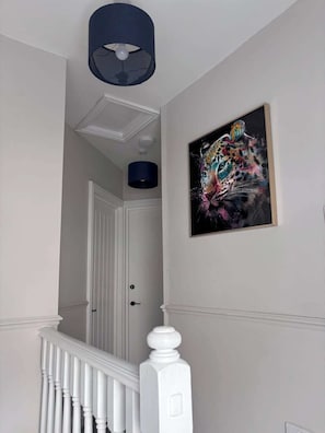 Een gang met een witte deur, een blauwe hanglamp en een lijst met een abstract schilderij aan de muur.
