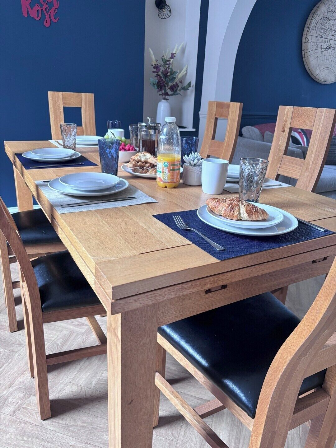 A dining table set with plates, cutlery, and a bottle of orange juice, surrounded by chairs with black cushions.