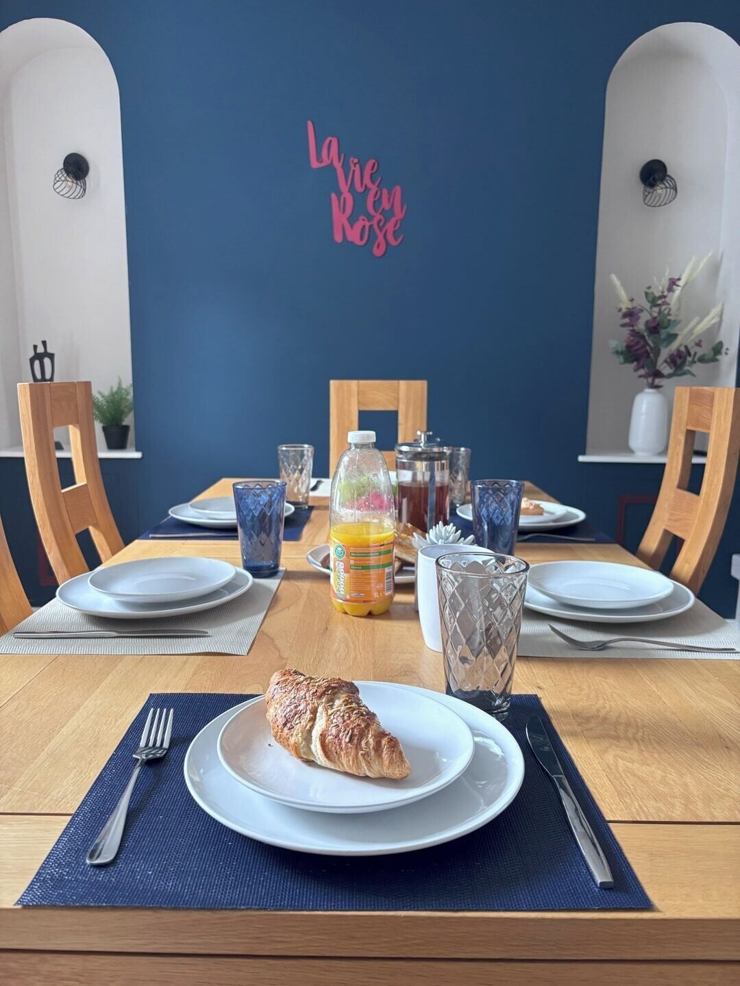 A dining table set with plates, cutlery, a bottle of orange juice, a glass, and a croissant.