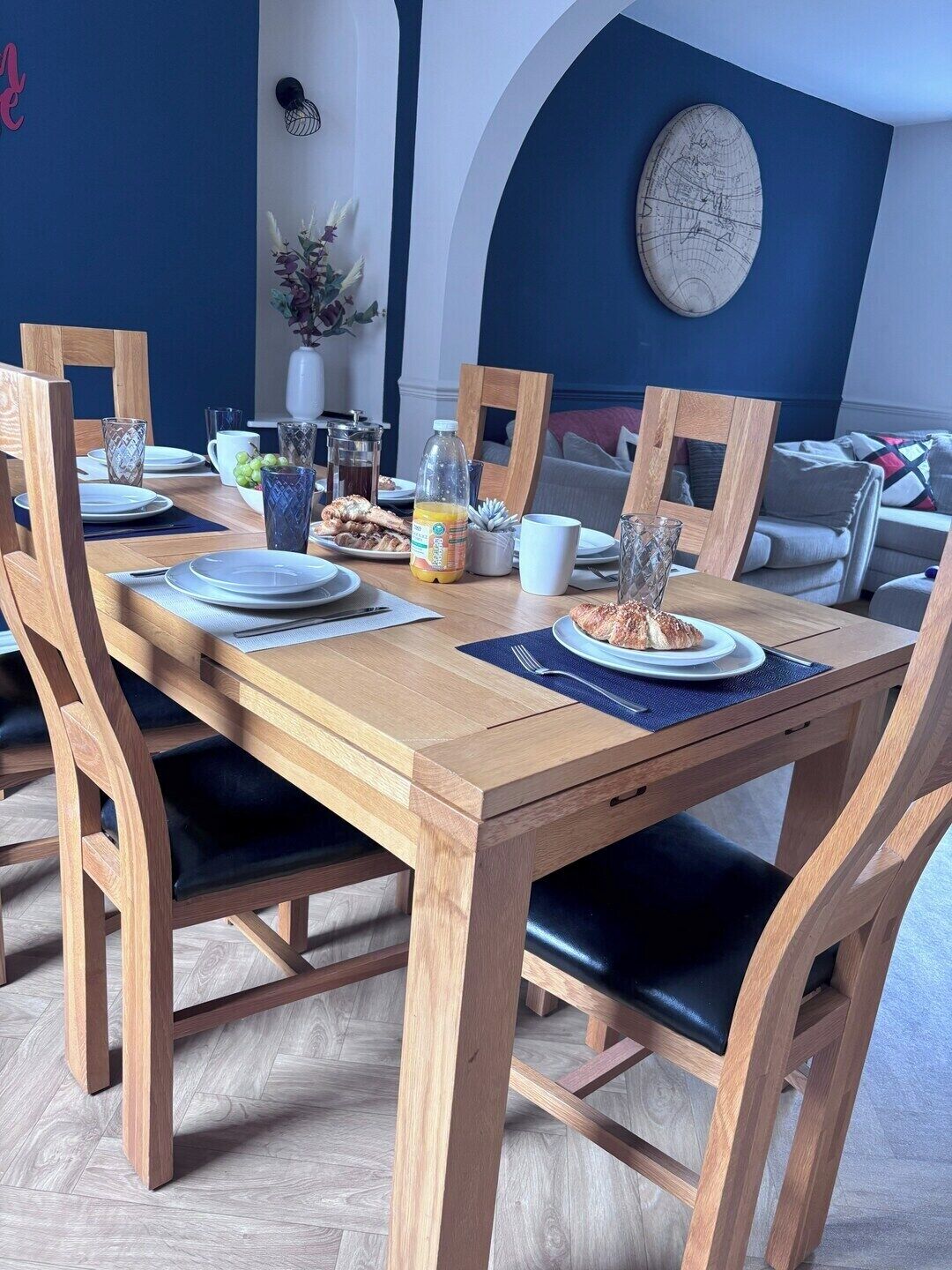 A dining area with a wooden table set for four, surrounded by chairs with black cushions.