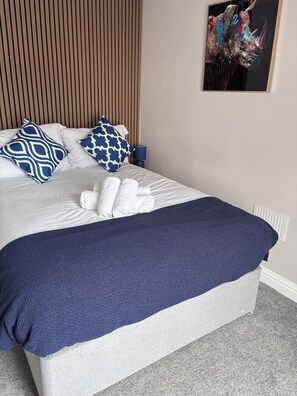 Cama bem arrumada com almofada estampada em azul e branco, manta azul escura e toalha branca.