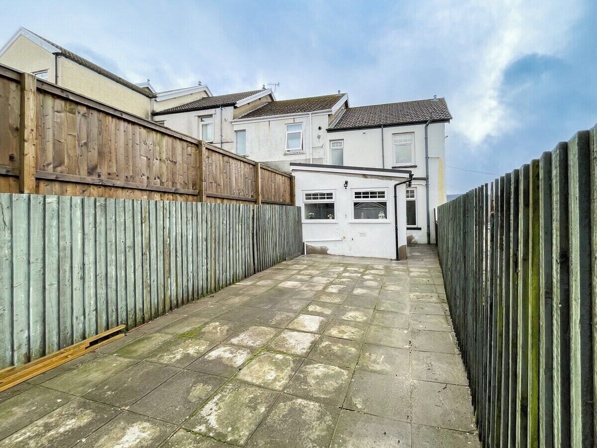A narrow paved pathway leading to a white building with a tiled roof, flanked by wooden fences.