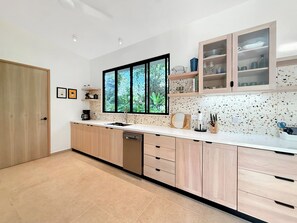 Une cuisine moderne équipée d’un lave-vaisselle, de meubles en bois, d’une grande fenêtre et d’un plan de travail sur lequel sont disposés divers objets.