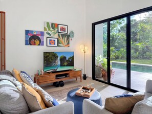 A modern living room with a flat-screen TV, a wooden entertainment unit, and a large sliding glass door leading to a pool. - Charming 3-bedroom house with AC in Playa Negra Playa Viejo (Playa Negra, Puerto Viejo de Talamanca)