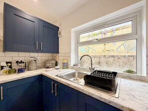 A kitchen with dark blue cabinets, a white countertop, a sink with a dish rack, and a window with a view of a wooden fence and plants.