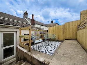 A patio area with a patterned rug, two chairs, a wooden fence, and a building with a tiled roof.