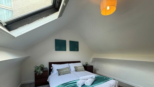 A bedroom with a skylight, a bed with a green bedspread, two wall-mounted artworks, and a nightstand with a plant.