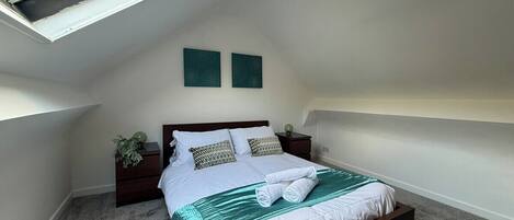 A bedroom with a skylight, a bed with a green bedspread, two wall-mounted artworks, and a nightstand with a plant.