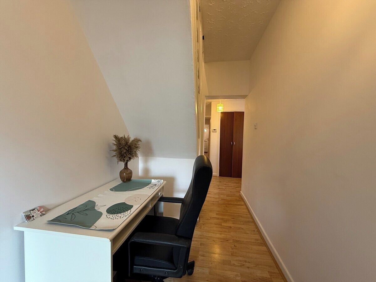A narrow hallway with a desk and chair, a vase with dried flowers, and a wooden floor.