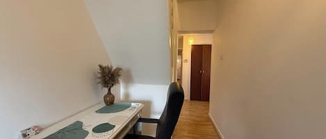 A narrow hallway with a desk and chair, a vase with dried flowers, and a wooden floor.