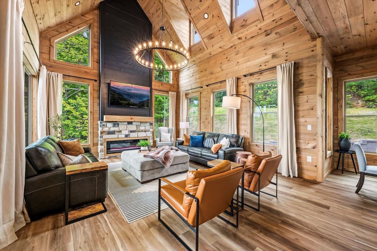 A spacious living room with a fireplace, wooden walls, and large windows.