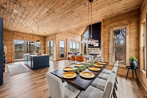 A spacious dining area with a long table set for a meal, wooden walls, and large windows. - Mtn Cabin w/ Hot Tub, Fire Pit & EV Charger (Sevierville)