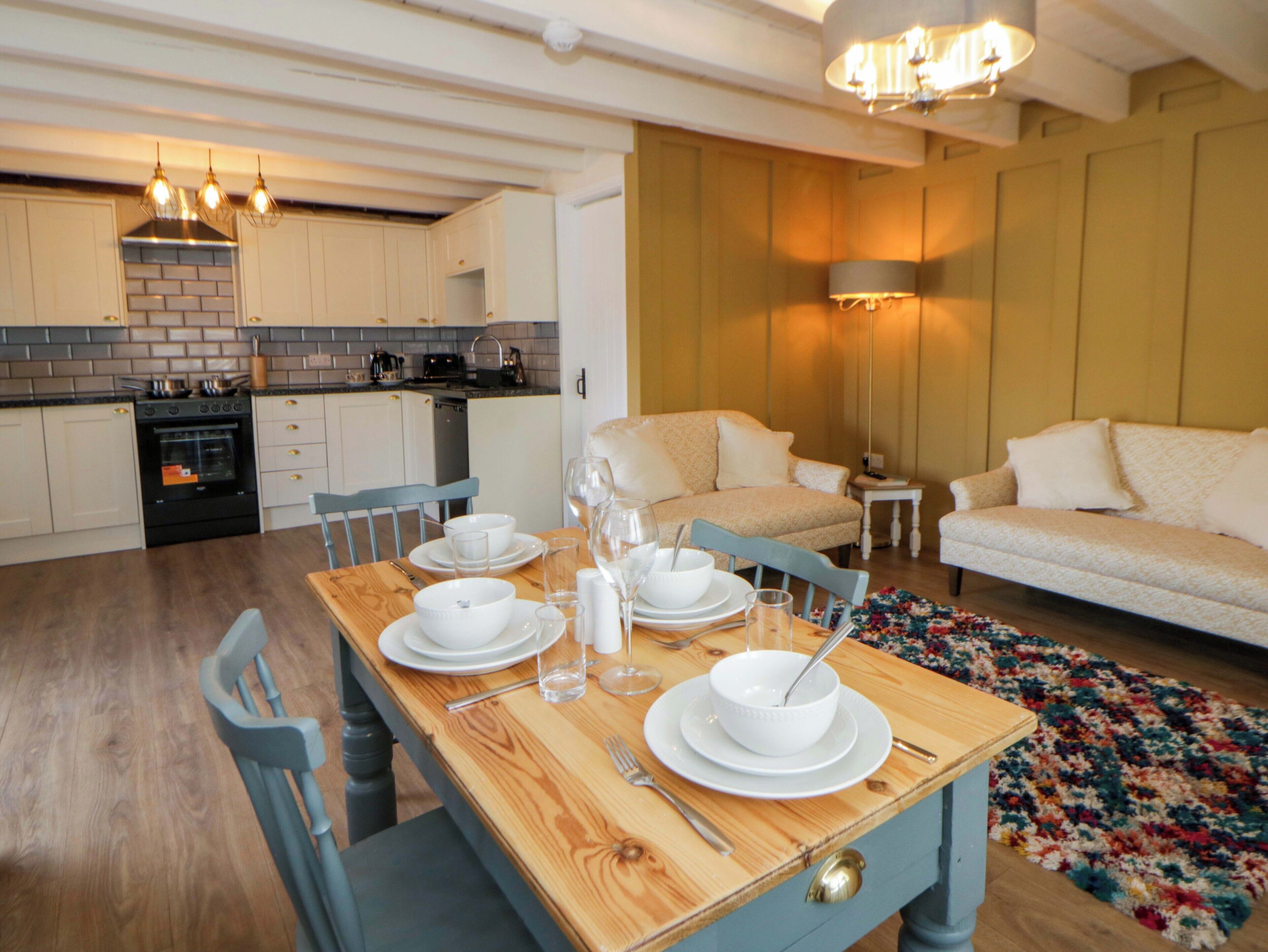 A dining area with a wooden table set for four, a kitchen in the background, and a living room with a sofa and a small table.