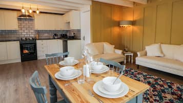 A dining area with a wooden table set for four, a kitchen in the background, and a living room with a sofa and a small table.