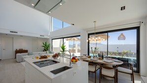 Una cocina moderna con una isla central, comedor con sillas de madera, ventanas amplias con vistas y un techo con iluminación empotrada.