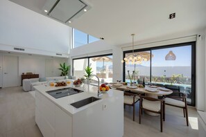 A modern kitchen with a central island, dining area with chairs, and large windows offering a view of the sea. - Luxury Pool Villa by Fidalsa (Polop)