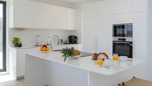 Una cocina moderna con muebles blancos, una isla central con fruta y productos para el desayuno, horno empotrado y cafetera.