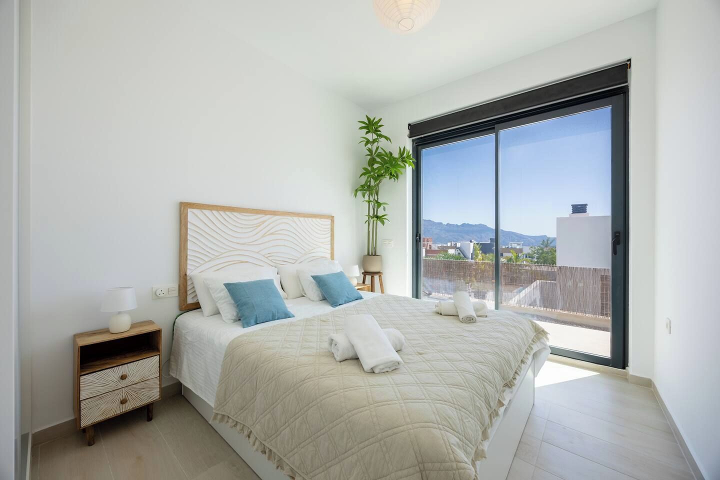Un dormitorio con cama, mesitas de noche, una planta en maceta y vistas a través de un ventanal corredera.