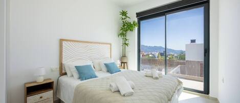 Un dormitorio con cama, mesitas de noche, una planta en maceta y vistas a través de un ventanal corredera.