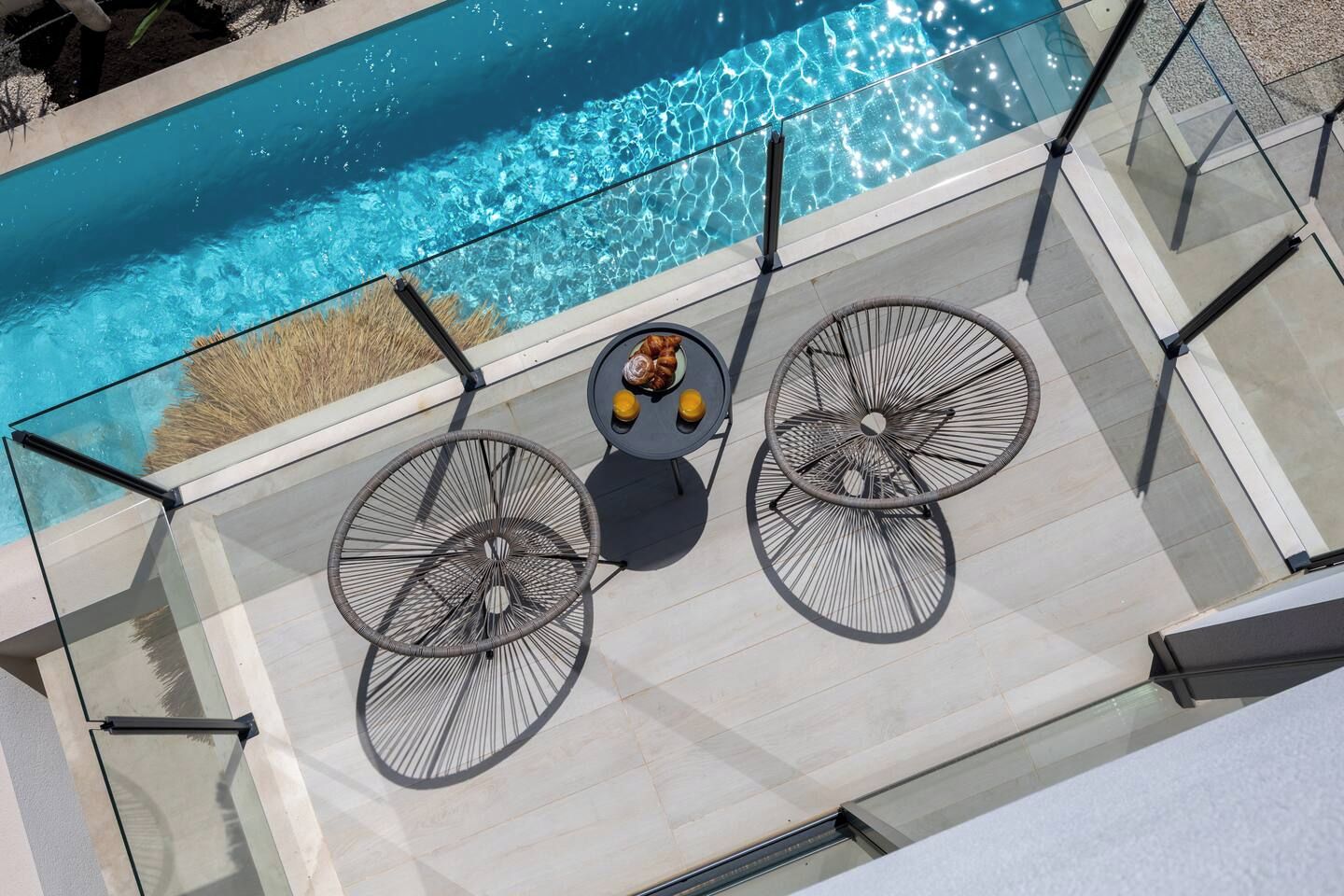A rooftop pool area with a glass balustrade, two chairs, and a table with a fruit bowl.